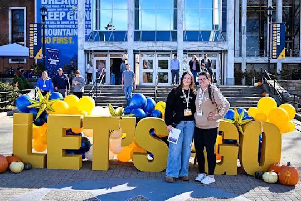 Two prospective students pose for a photo in front of the big Let's Go letters decorated with gold and blue balloons. The Mountainlair Student Union is in the background.